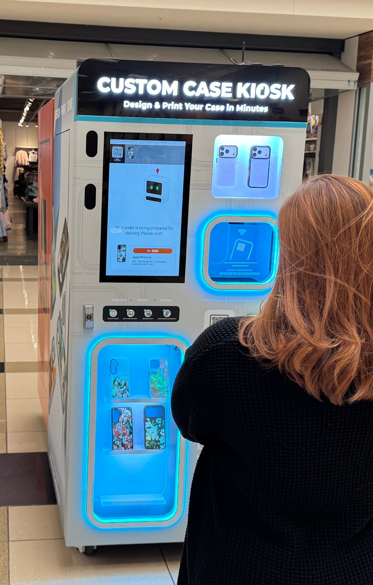 Custom Case Kiosk at Woodland Mall in Grand Rapids, Michigan — a shopper at the touchscreen while the kiosk prints a case, backlit display shelves alongside